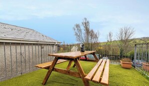 A wooden picnic table with benches on a grassy lawn, a metal fence, and a building with a sloped roof in the background. - 3BR House, Central, Pet Friendly, Next to Café (Ballycastle)