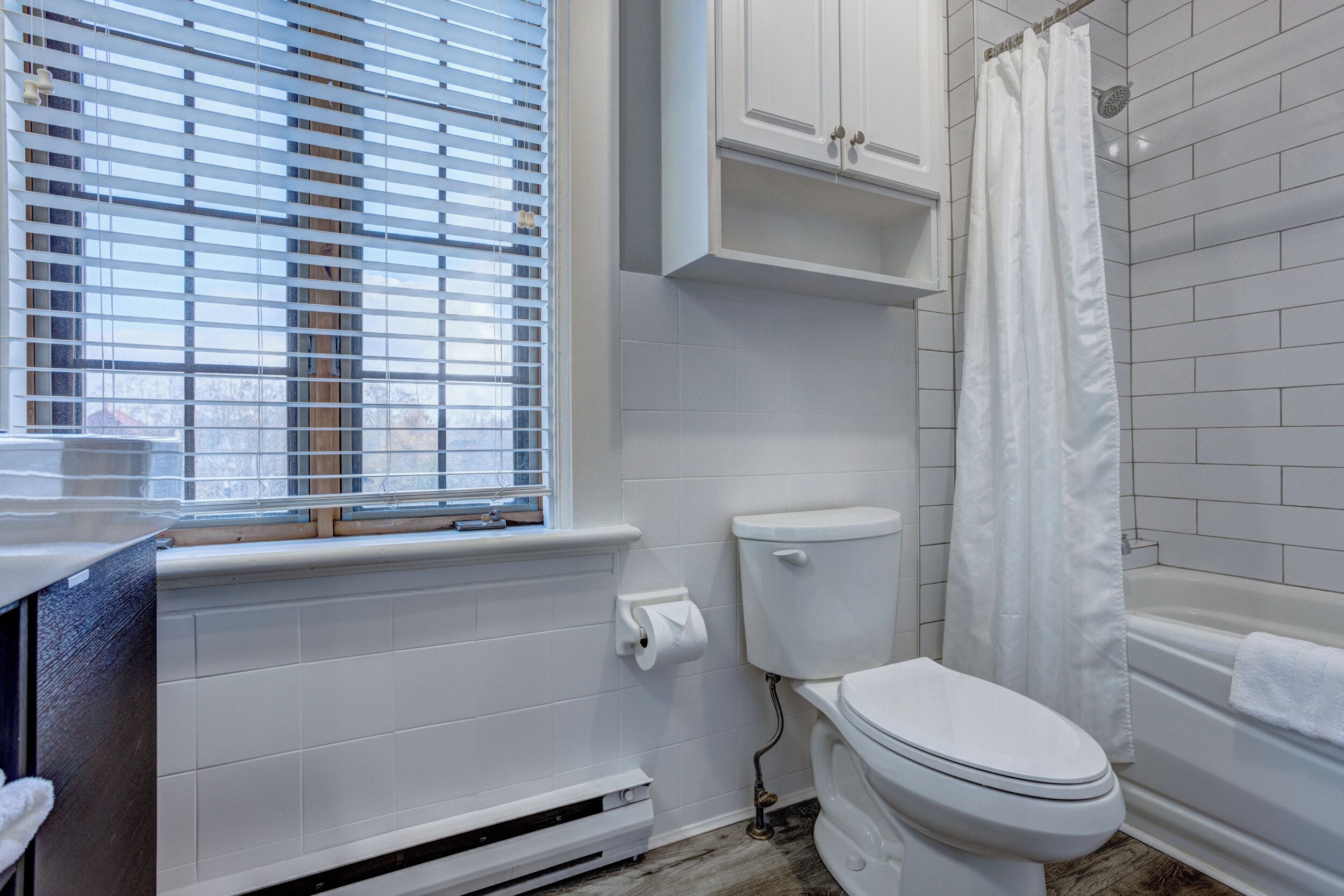 Um banheiro com vaso sanitário, banheira com cortina de chuveiro, janela com persianas e parede revestida de azulejos brancos.