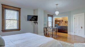 A bedroom with a large bed, a desk, and a kitchenette in the background.