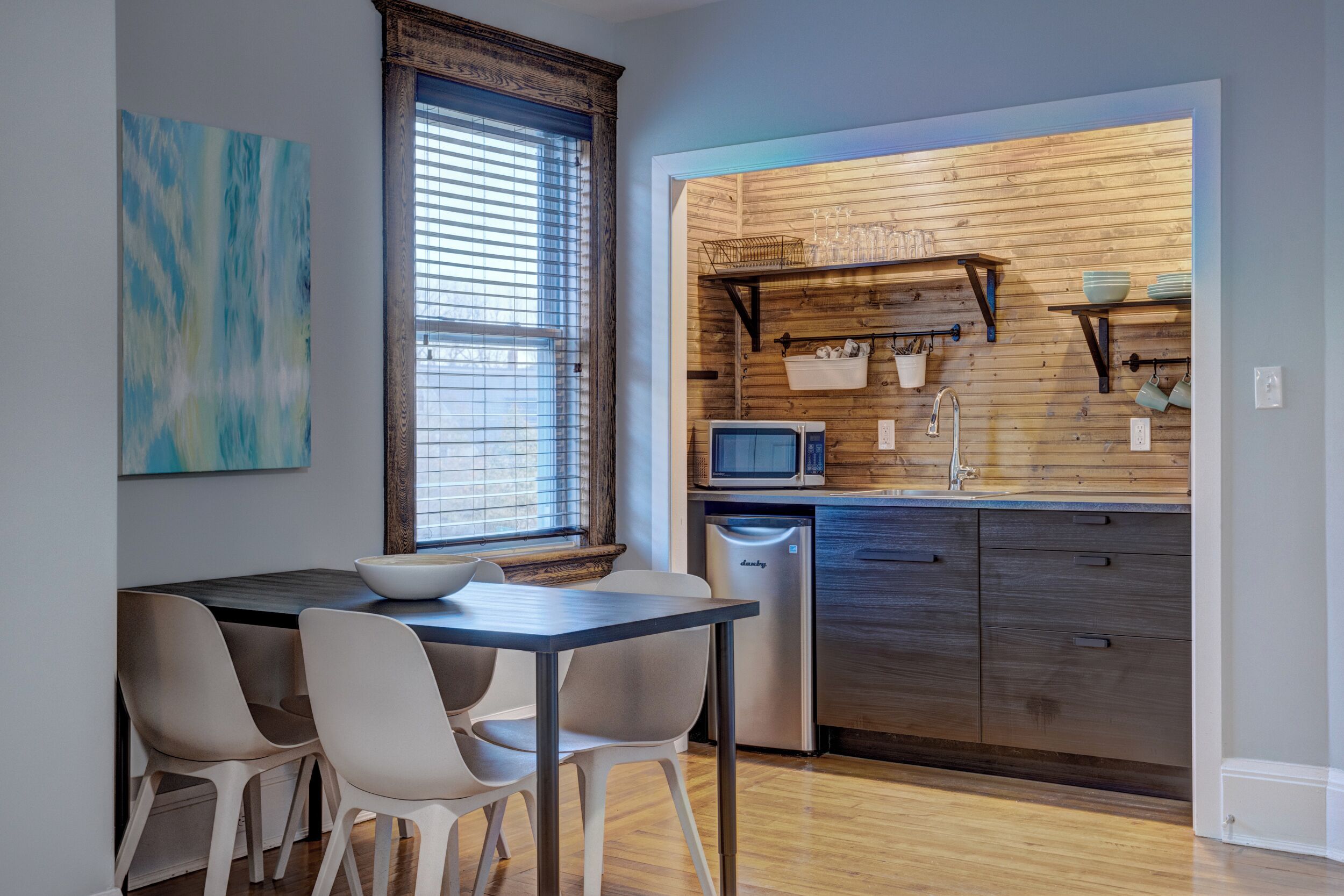 A modern kitchen with wooden paneling, a microwave, and a sink. There is a dining table with chairs and a window with blinds.