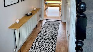 A hallway with framed artwork, a radiator, a wooden bench, and a patterned rug leading to an outdoor area with a table and chairs. - Quiet 2BR Home with Garden, Sleeps 5 (Londonderry)