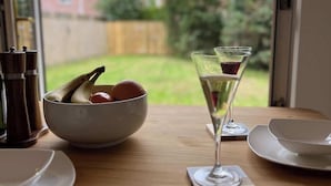 A dining table with a bowl of fruit, a glass of wine, and a view of a backyard through a window. - Quiet 2BR Home with Garden, Sleeps 5 (Londonderry)