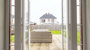 Eine Terrasse mit einem Rattan-Sofa, einer Glastür, die nach draußen führt, und Blick auf ein Haus gegenüber dem Garten.