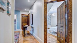 A hallway with wooden flooring, a dining area with a table and chairs, and a bedroom with a bed and a chandelier. - Executive 1-Bedroom King Bed Suite in Lunenburg (Suite 5) (Lunenburg)