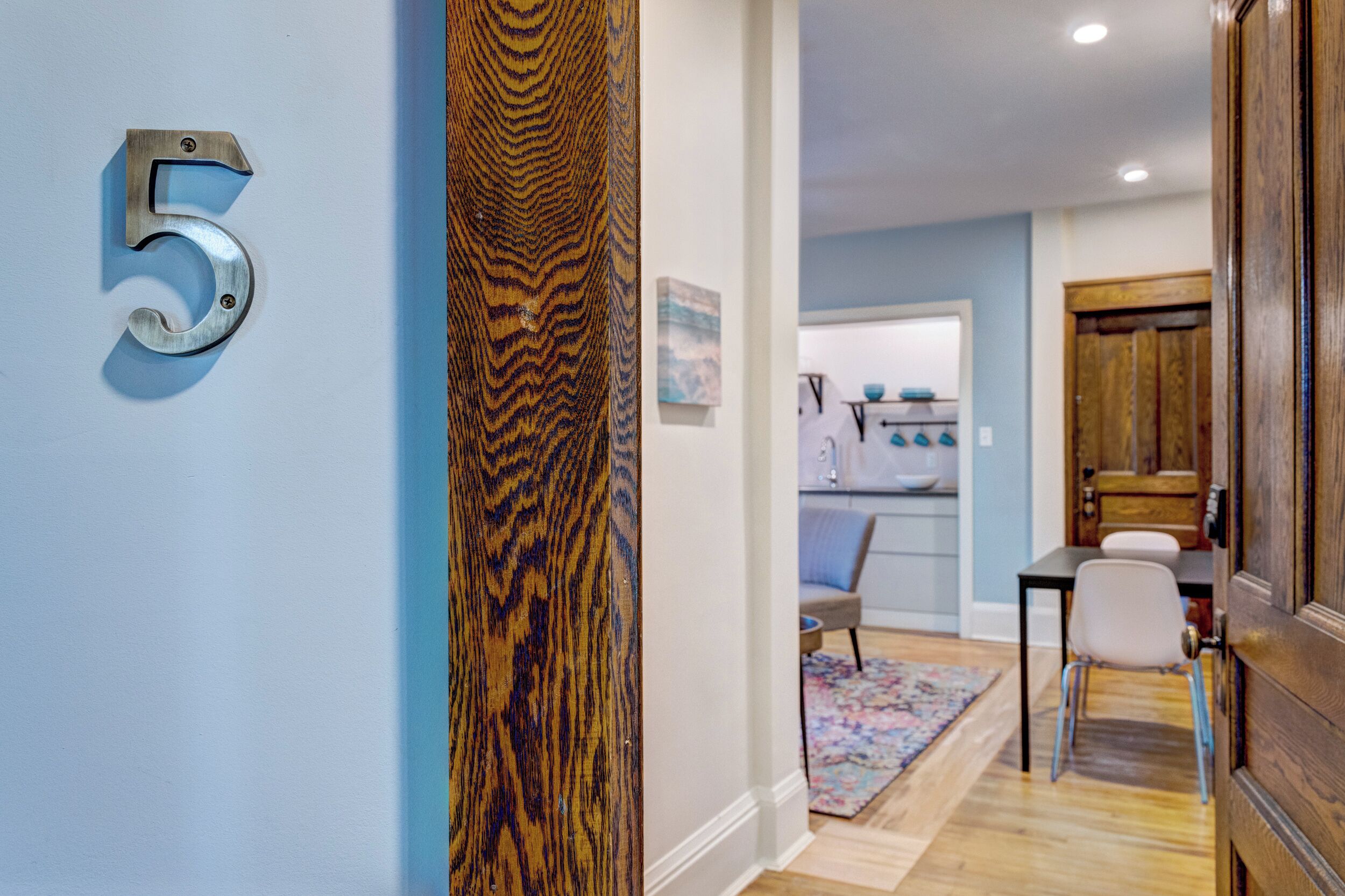 A hallway with a number '5' sign, a wooden door, a dining area with a table and chairs, and a kitchen in the background.