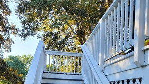 A white wooden staircase with black steps leading up to a white railing, surrounded by greenery.