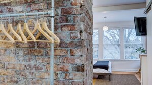 A room with a brick wall, wooden hangers, a bed, a TV, and a window with blinds.