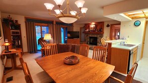 Een gezellige woonkamer met een houten eettafel, comfortabele zitplaatsen, een open haard, een televisie en een keuken met een spoelbak en aanrecht.