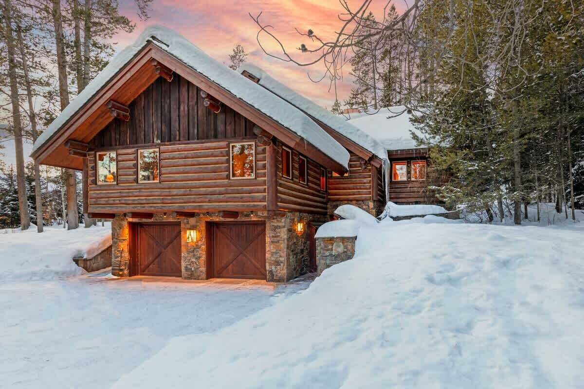 A log cabin with a stone foundation, large wooden doors, and a snow-covered roof.