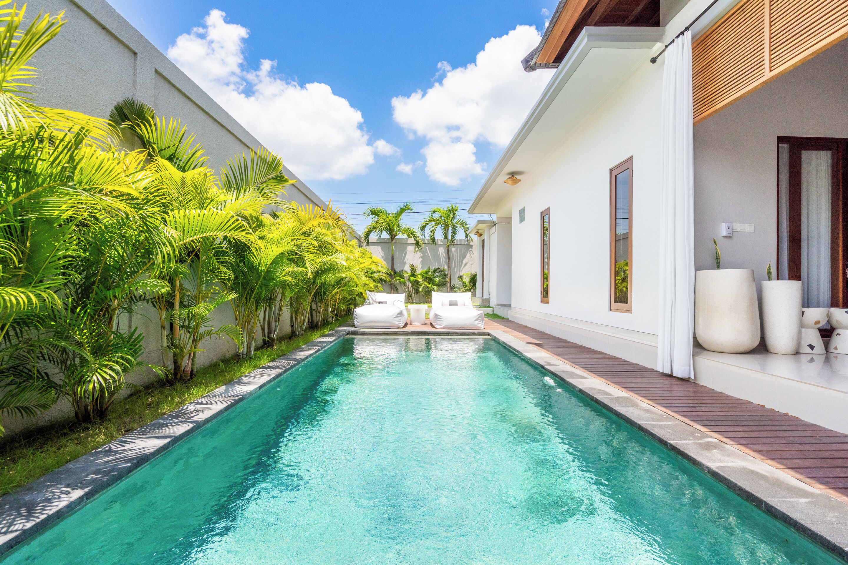 A swimming pool with two lounge chairs, surrounded by lush greenery and a white building with a wooden deck.