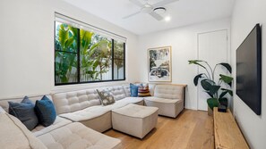 A modern living room with a sectional sofa, a ceiling fan, a large window with a view of greenery, and a mounted TV. - Cylinders Coastal Deluxe (Kingscliff)