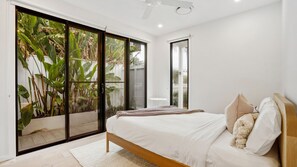 Un dormitorio con un amplio ventanal corredera que da a un balcón, una cama con ropa de cama blanca y una pequeña mesa blanca.