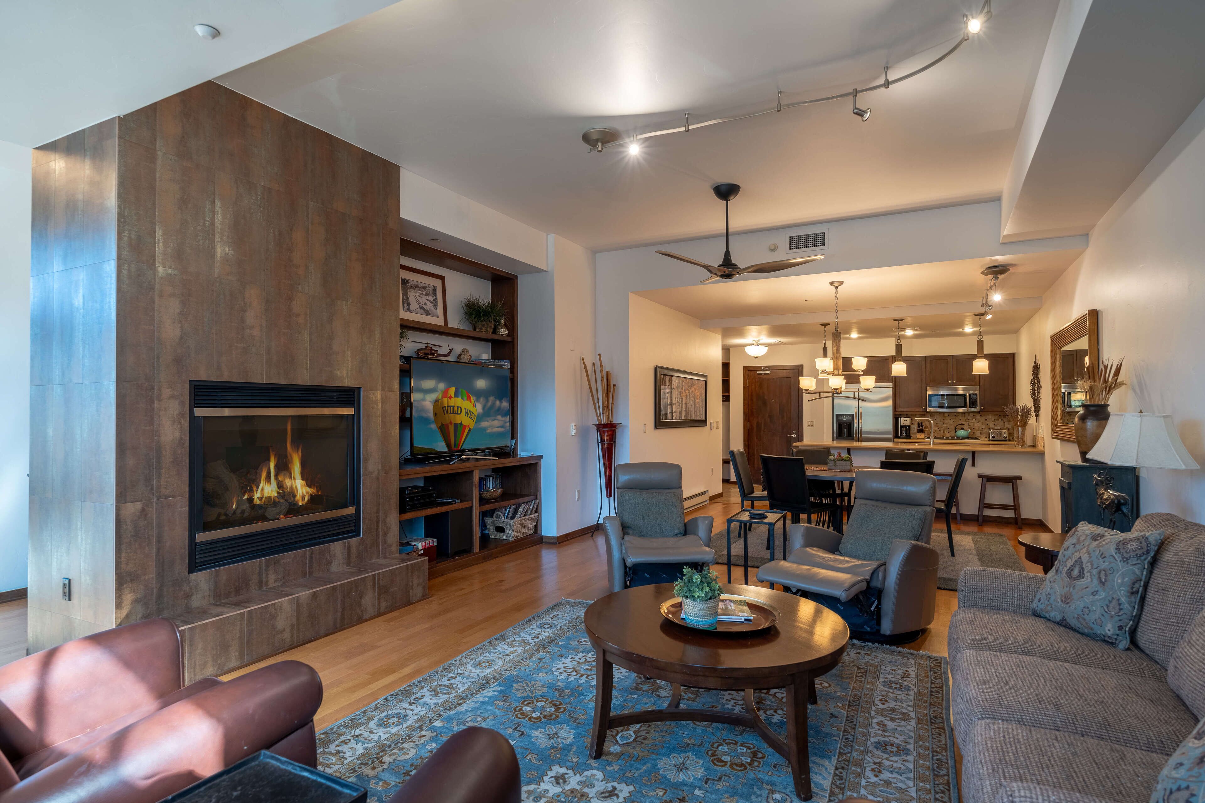 A spacious living room with a fireplace, comfortable seating, and a kitchen in the background.