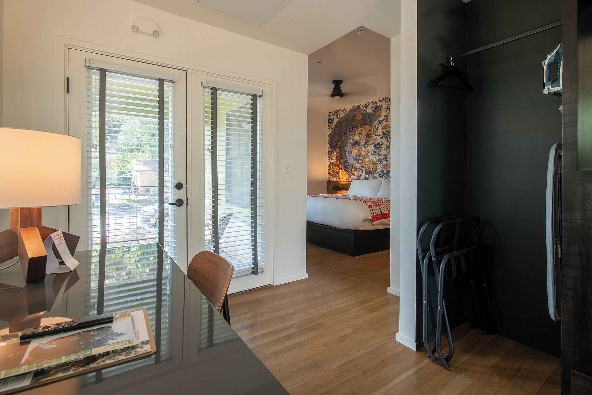 Une chambre d’hôtel moderne dotée d’une porte vitrée donnant sur un balcon, d’un lit avec une tête de lit à motifs floraux et d’un mur noir orné d’un fer à repasser suspendu.