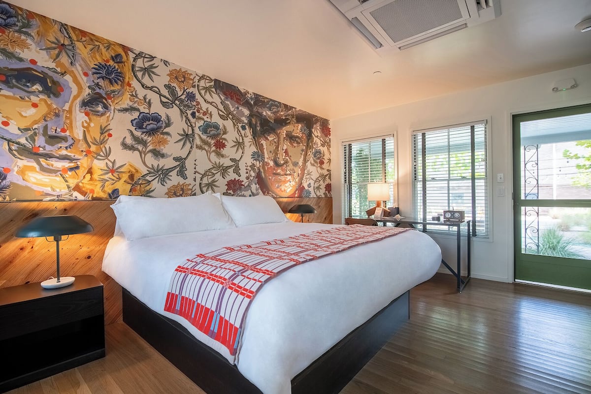Un dormitorio moderno con una cama grande, un mural floral, una mesita de noche con lámpara y vistas a zonas verdes por la ventana.