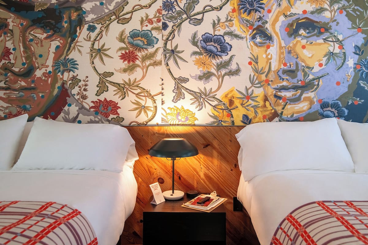 Una habitación de hotel con dos camas, una mesita de noche con lámpara y un tapiz decorativo en la pared.