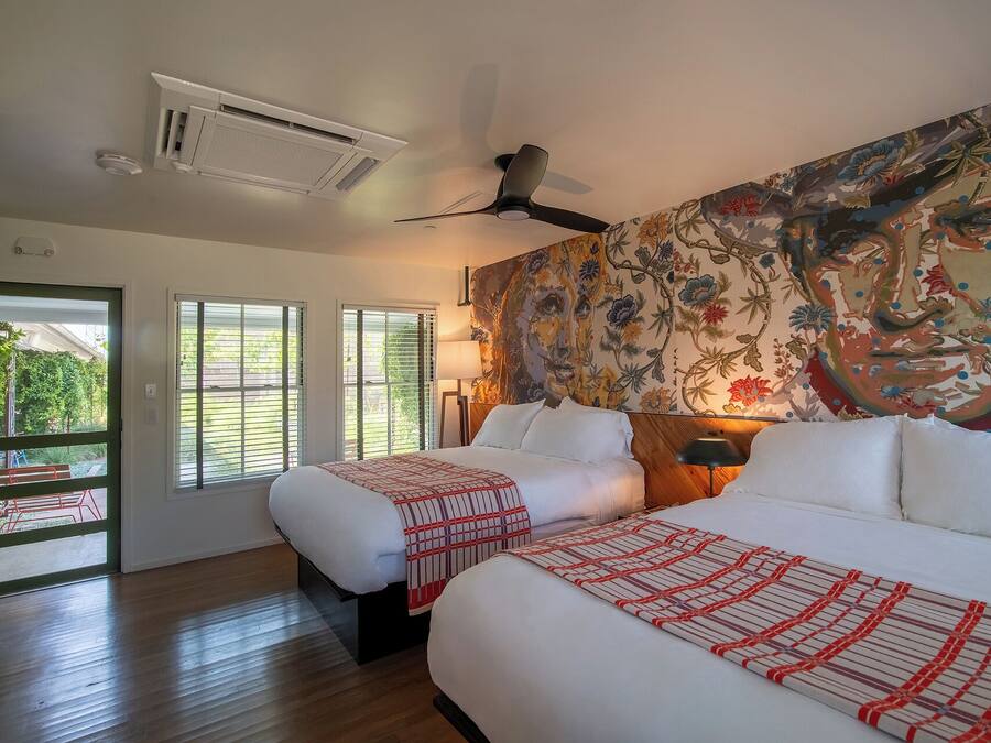 A hotel room with two beds, a ceiling fan, a large floral mural, and a view of the outdoors through the windows.