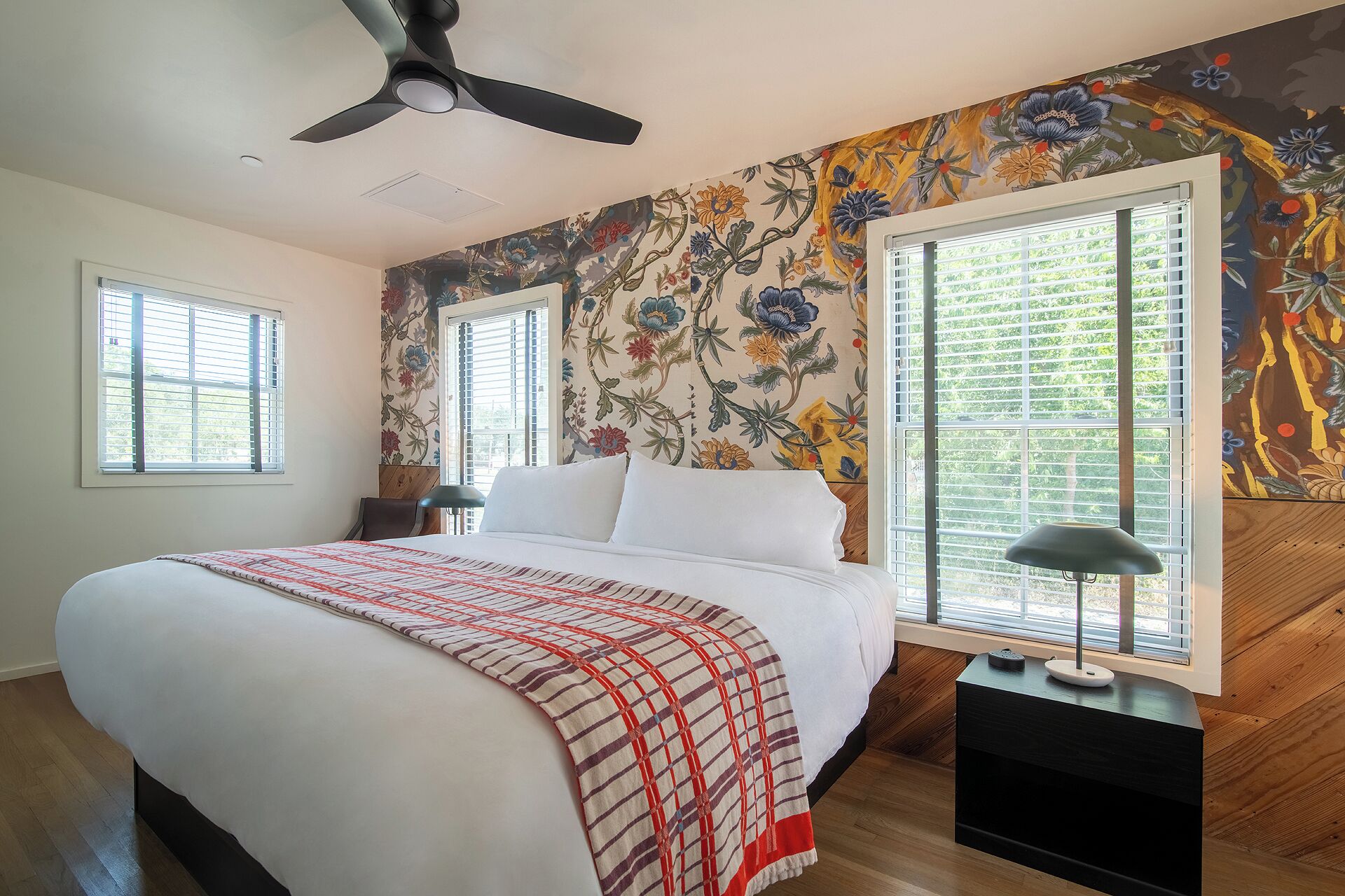 Quarto com cama, duas janelas com persianas, ventilador de teto e papel de parede floral.