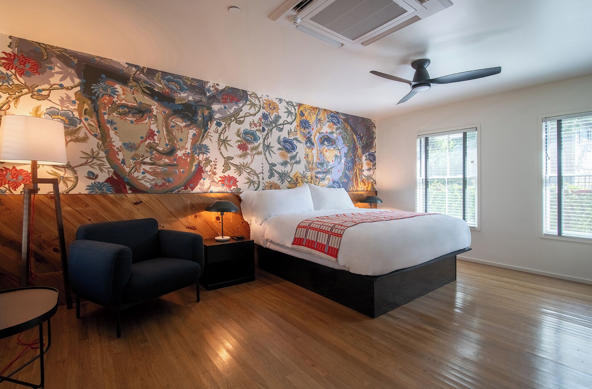 Una habitación de hotel moderna con una cama grande, cabecera de madera, un escritorio con lámpara, una silla y un tapiz floral grande en la pared.
