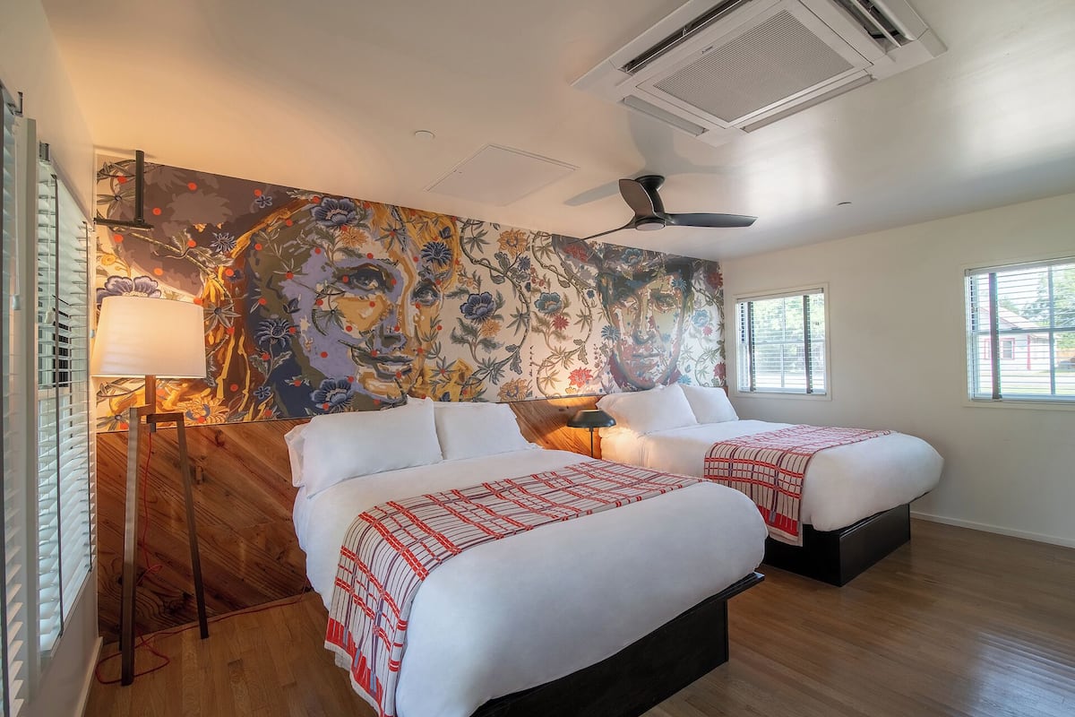 Habitación de hotel con dos camas, un mural floral grande, un ventilador de techo y una ventana con persianas.