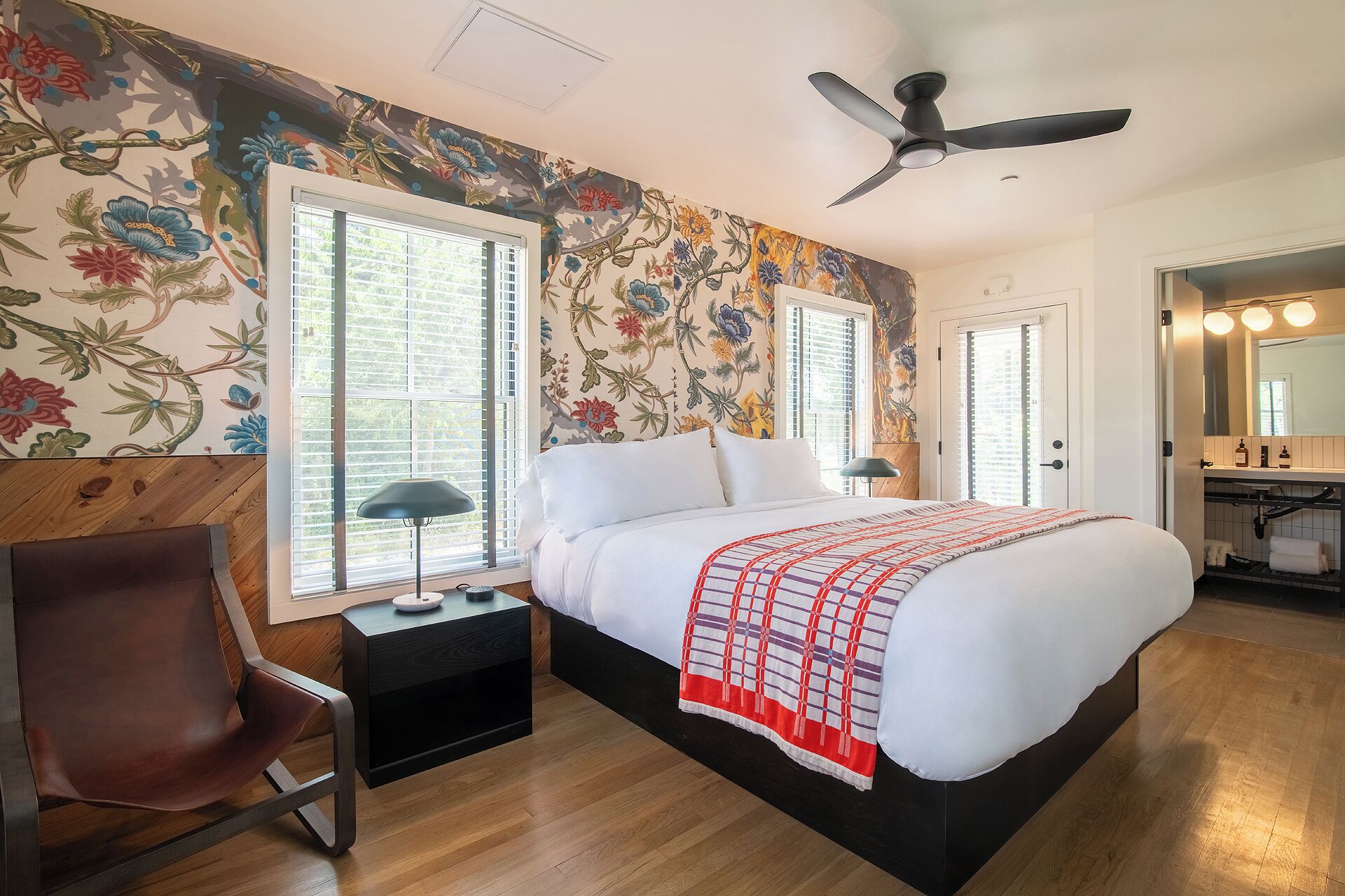 A bedroom with a bed, a chair, a nightstand, a ceiling fan, and a bathroom with a mirror and lights.