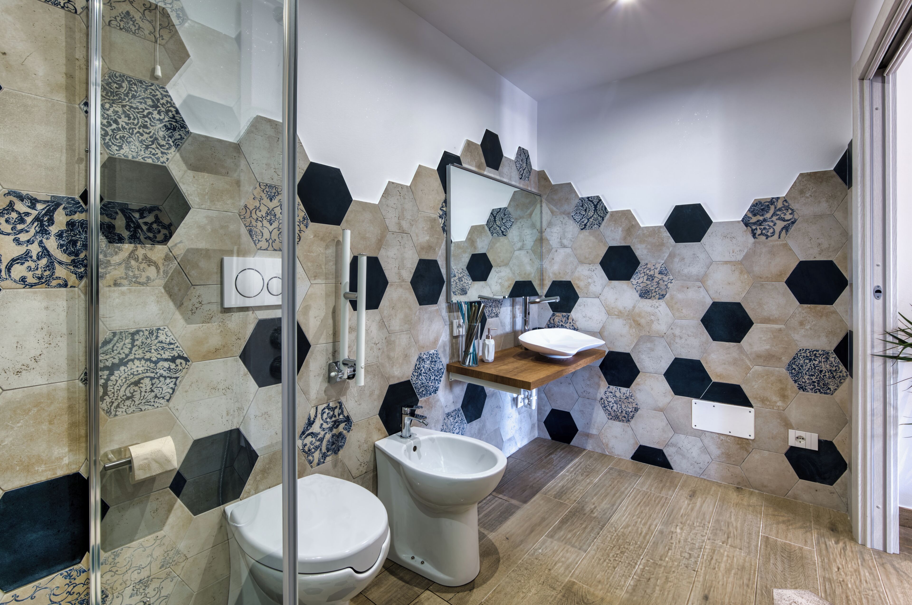 Une salle de bain moderne avec un mur recouvert de carreaux hexagonaux, une douche en verre, des toilettes et un lavabo doté d’un plan de travail en bois.