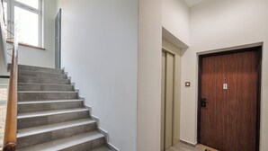 Un escalier menant à une porte avec une plaque numérotée, un paillasson et une fenêtre donnant sur des arbres.