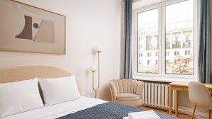 Une chambre d’hôtel équipée d’un lit, d’un bureau, d’une chaise, d’un radiateur et d’une fenêtre donnant sur des immeubles.