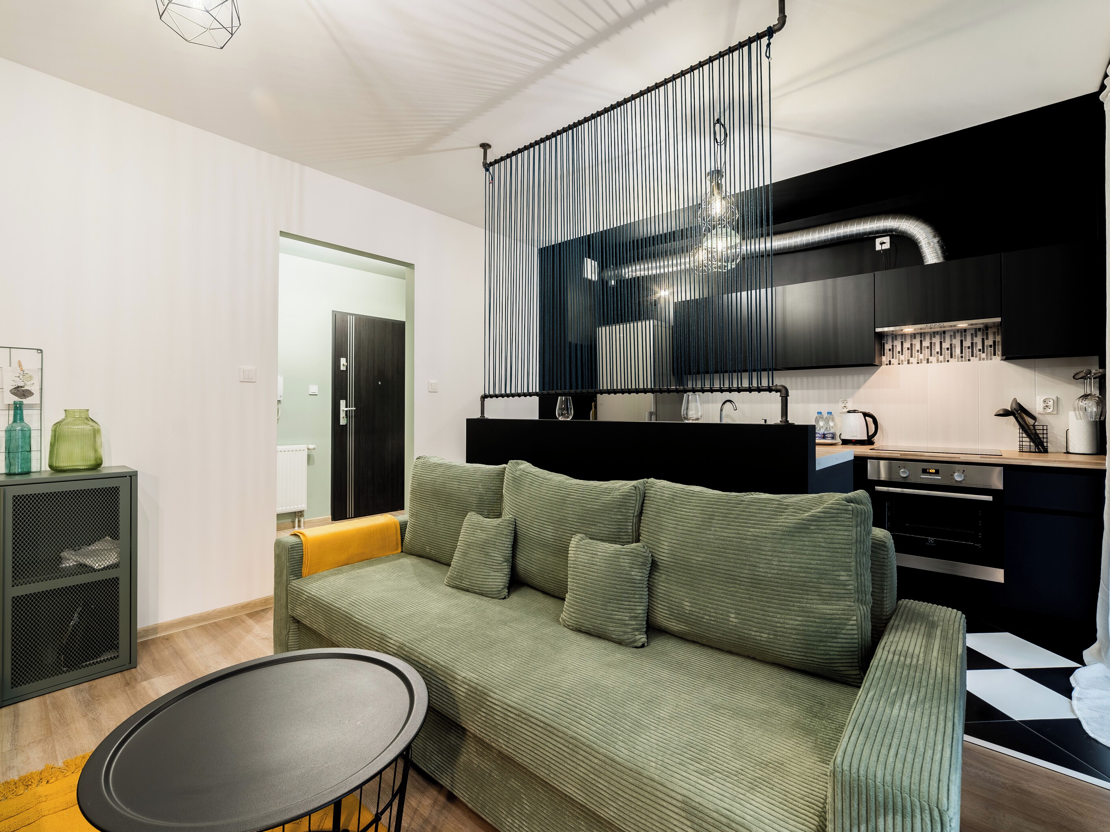 Una sala de estar moderna con un sofá verde, una mesita redonda y una zona de cocina con encimera negra y electrodomésticos de acero inoxidable.