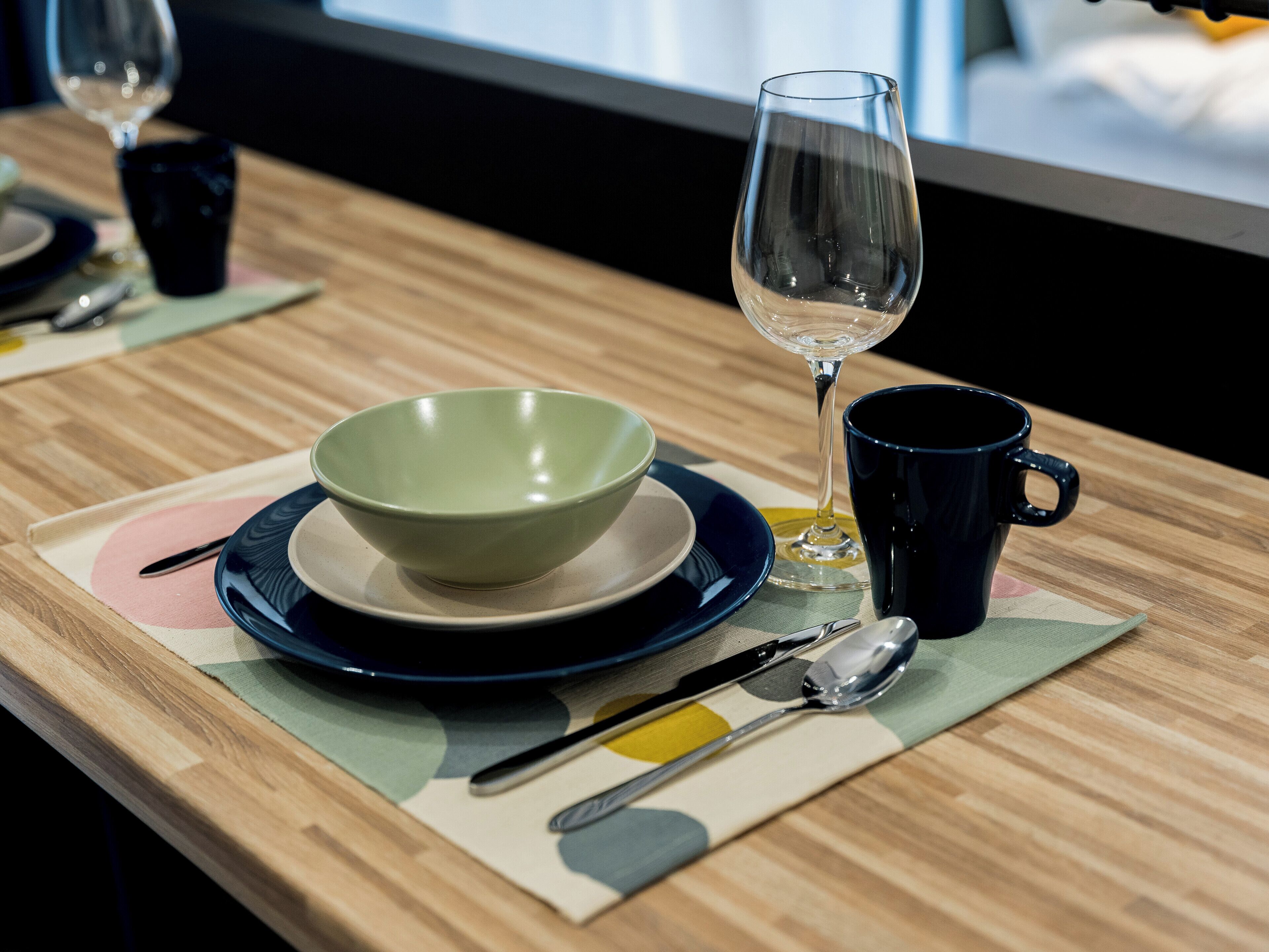Een tafel gedekt met een groene kom, een wijnglas, een zwarte mok, een lepel en een mes op een houten tafel met een kleurrijke placemat.
