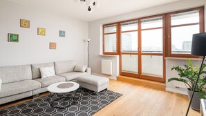 Una sala moderna con un sofá seccional gris, una mesa de centro redonda y un tapete con estampado blanco y negro. Hay varios cuadros enmarcados en la pared, una lámpara de pie y un ventanal con vistas a la ciudad.