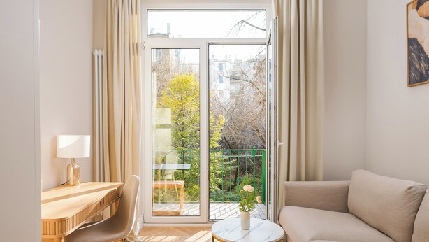A modern living room with a sofa, a small round table with a vase, a desk with a chair, and a sliding glass door leading to a balcony with a green railing and a tree. - Urban Heart Deluxe Studio #2 (Warszawa)