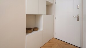 A hallway with white walls, a wooden floor, and a white door with a silver handle. There are two woven baskets on a shelf. - Urban Heart Deluxe Studio #1 (Warszawa)
