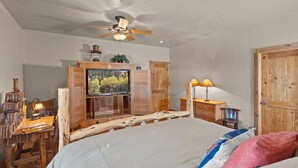 A bedroom with a wooden bed, a television mounted on a wooden cabinet, a ceiling fan, and a wooden door.