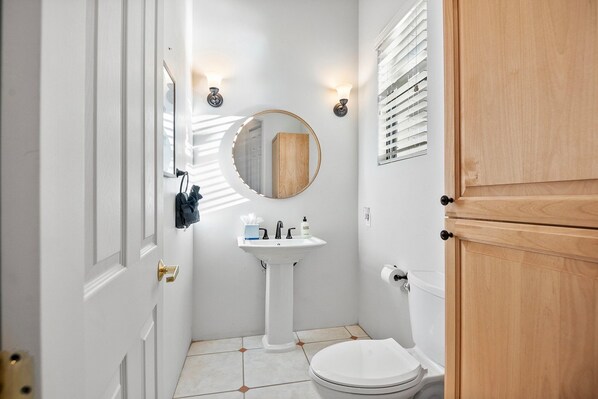 Une petite salle de bain blanche, avec un lavabo sur colonne, un miroir rond et des toilettes.