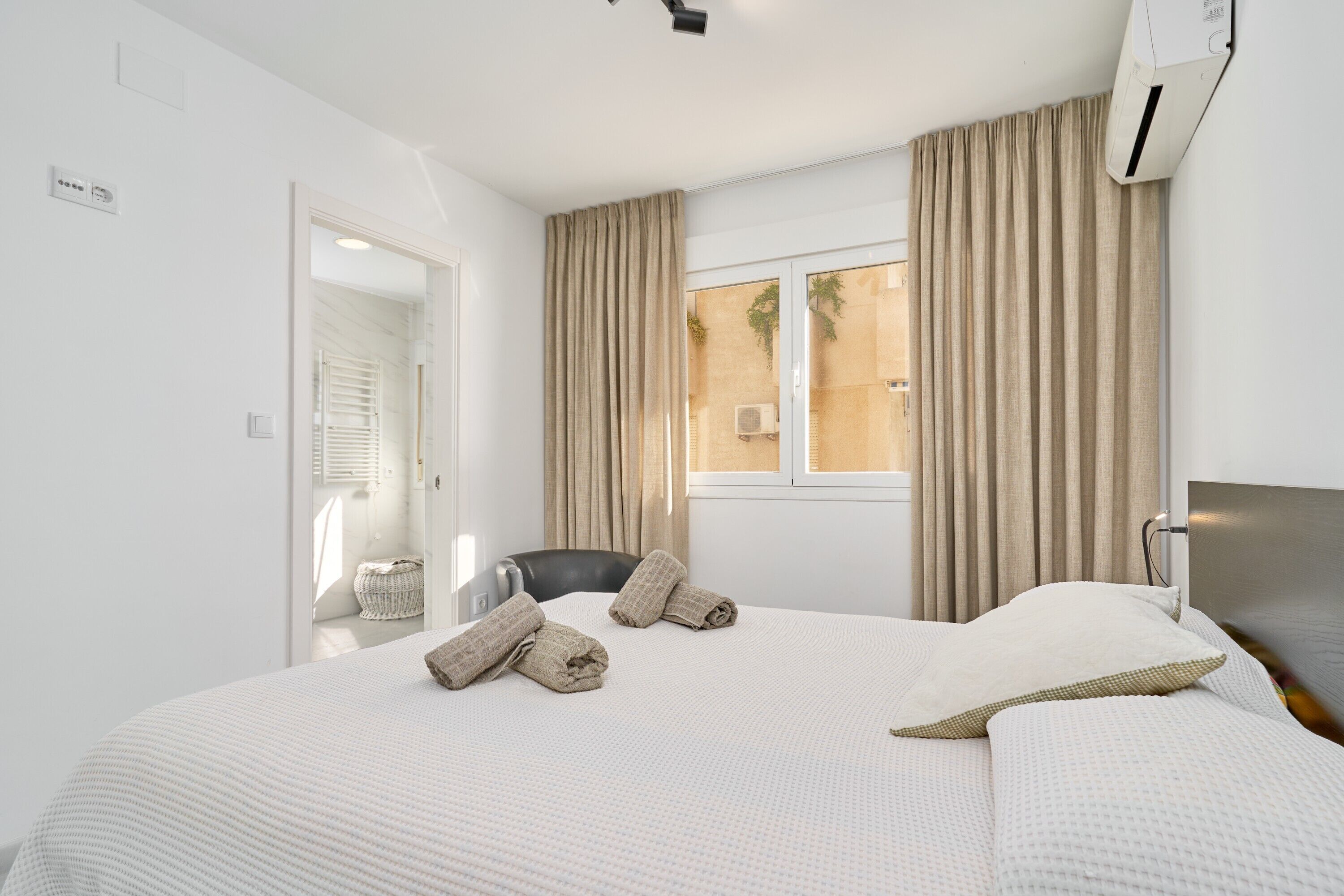 A hotel room with a bed, a chair, a window with curtains, and an air conditioning unit.