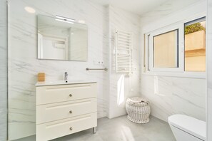 A bathroom with a white marble wall, a white sink with a mirror above it, a white toilet, and a wicker basket on the floor. - Apartment 'Beachfront Los Novos La Herradura' with Sea View, Wi-Fi and Air Conditioning (La Herradura)