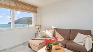 Un salon avec un canapé, une table basse ornée d’un vase de fleurs, une télécommande et une fenêtre donnant sur la mer et une montagne.