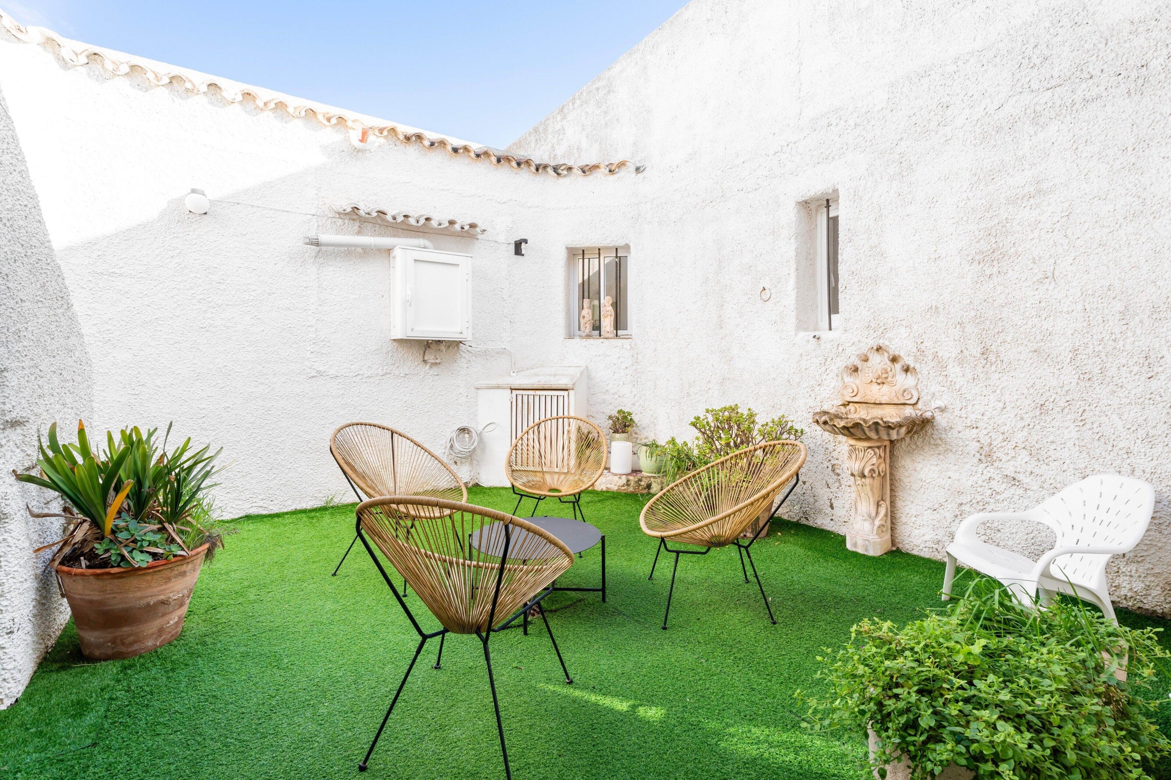 Un patio con césped artificial verde, sillas de mimbre, una pared blanca y una fuente de piedra.