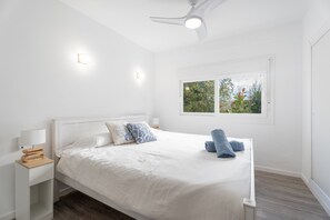 Ett sovrum med en säng, sängbord, en takfläkt och ett fönster med utsikt över grönområden.