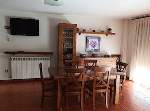 A dining area with a table and chairs, a cabinet with glass doors, a television mounted on the wall, and a radiator below.