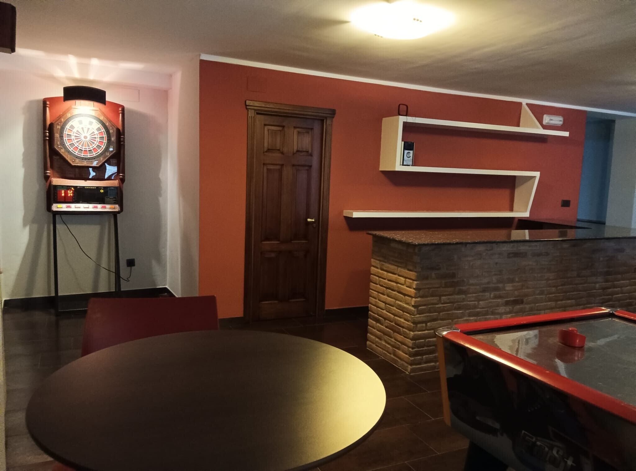 A room with a dartboard, a door, a brick fireplace, and a shelf.