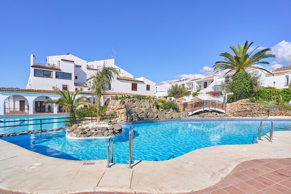 A swimming pool with a stone island, white buildings with balconies, and a clear blue sky.