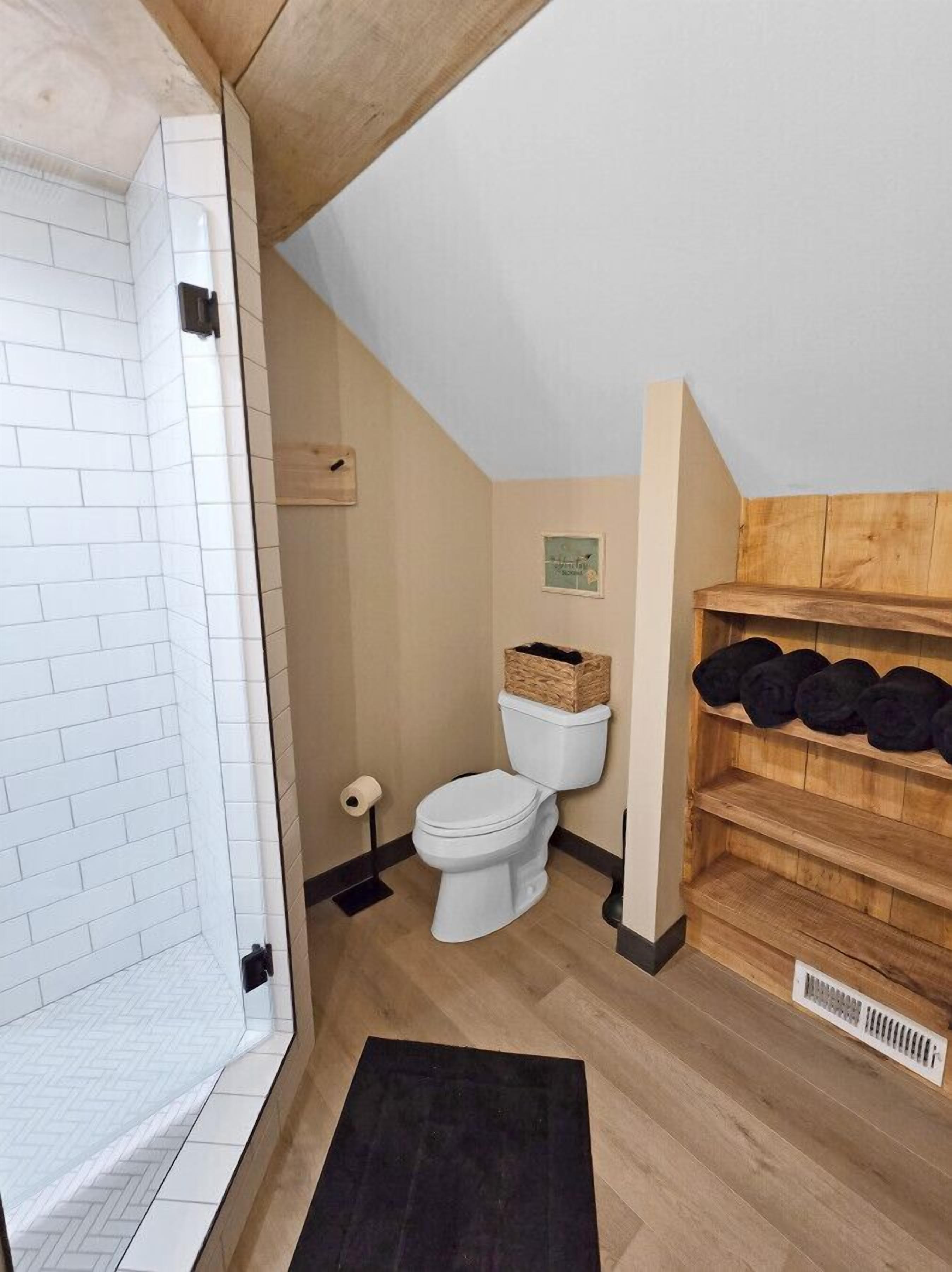 Ein Badezimmer mit einer weißen Toilette, einem Holzregal mit schwarzen Gegenständen, einem schwarzen Teppich und einem Fischgrätenparkettboden.