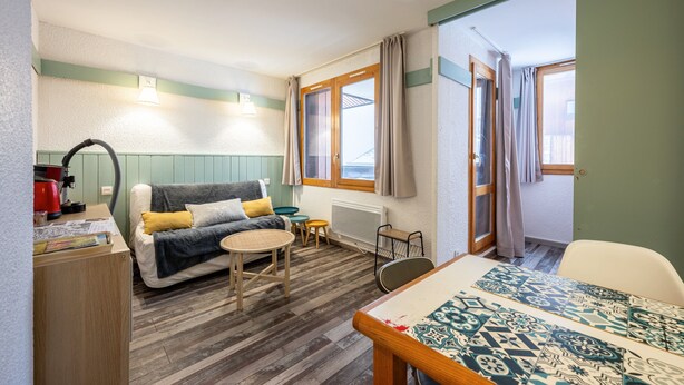 Una habitación compacta con sofá, una mesita, una mesa de comedor, un radiador y una ventana con cortinas.