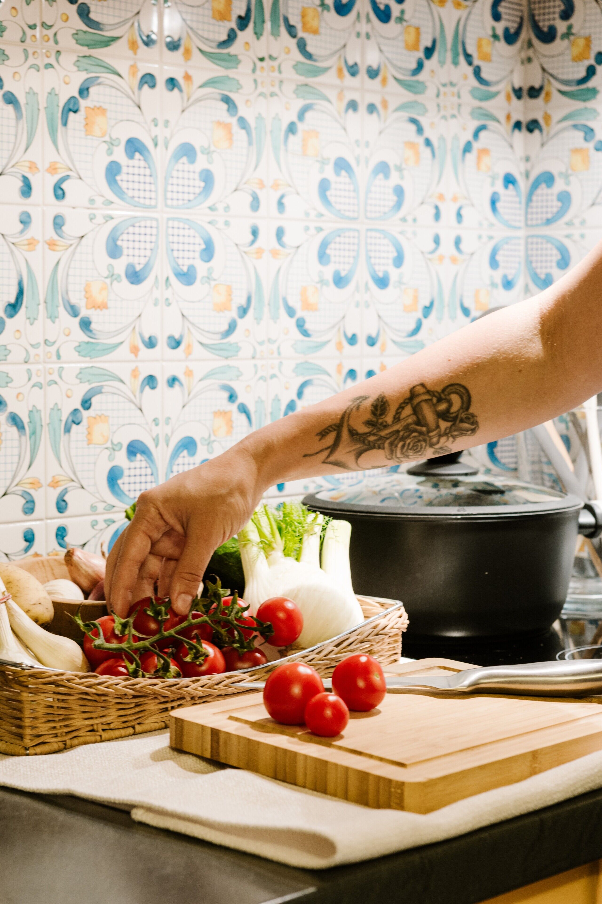 A kitchen with a patterned backsplash, a cutting board with tomatoes, a pot on the stove, and a basket of vegetables.