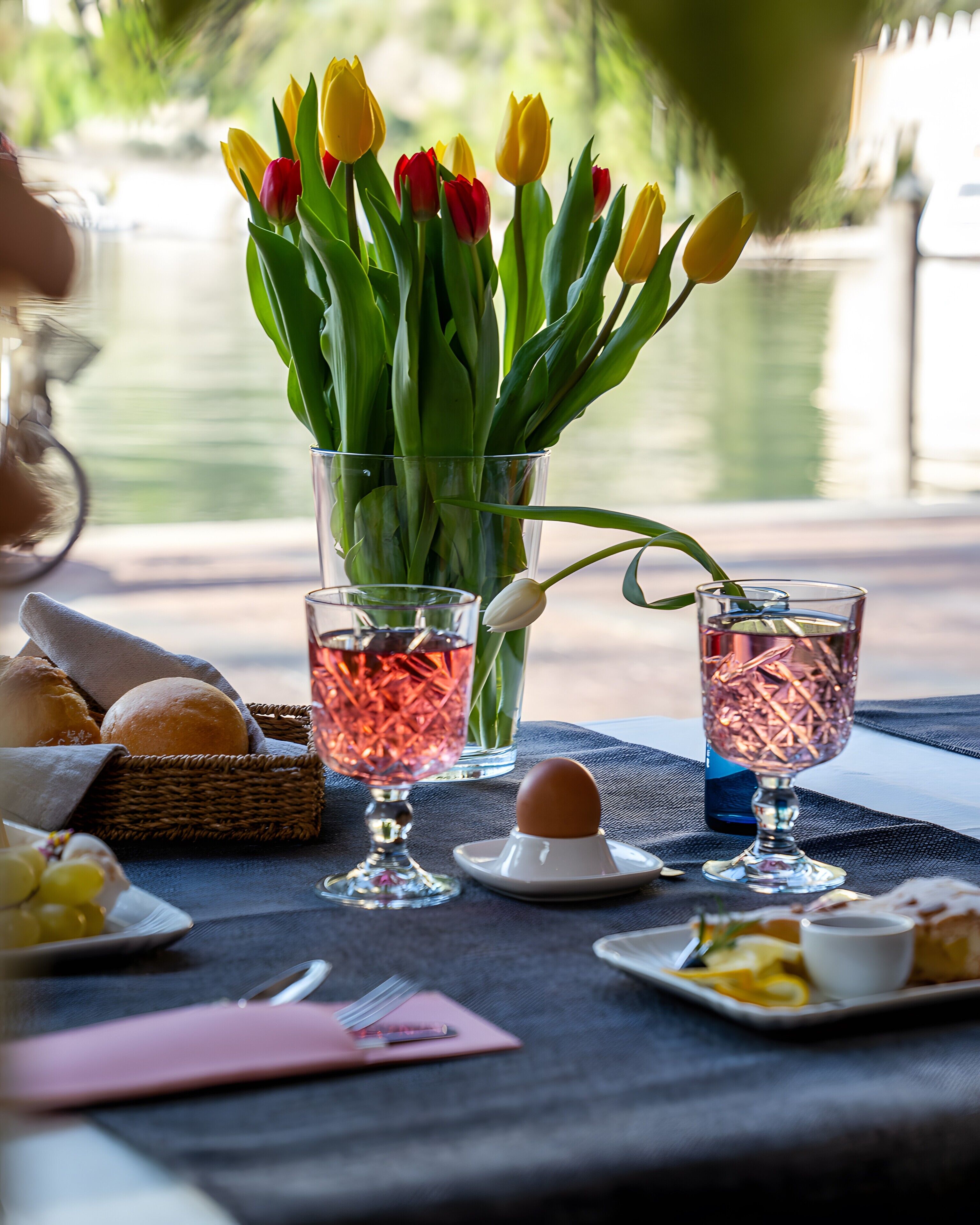Ein Tisch mit einer Glasvase voller Tulpen, zwei Gläsern mit roten und blauen Flüssigkeiten, einem Brotkorb, einer Traubenteller und einem Eierchen auf einer Serviette.