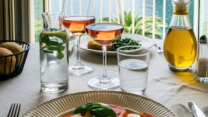 A table with a plate of food, glasses, and a bottle, with a scenic view of a mountain and sea through a window.