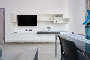 Un salon moderne doté d’un téléviseur à écran plat fixé au mur, d’une table à manger avec des chaises et d’un petit coin cuisine avec des meubles et des étagères blancs.