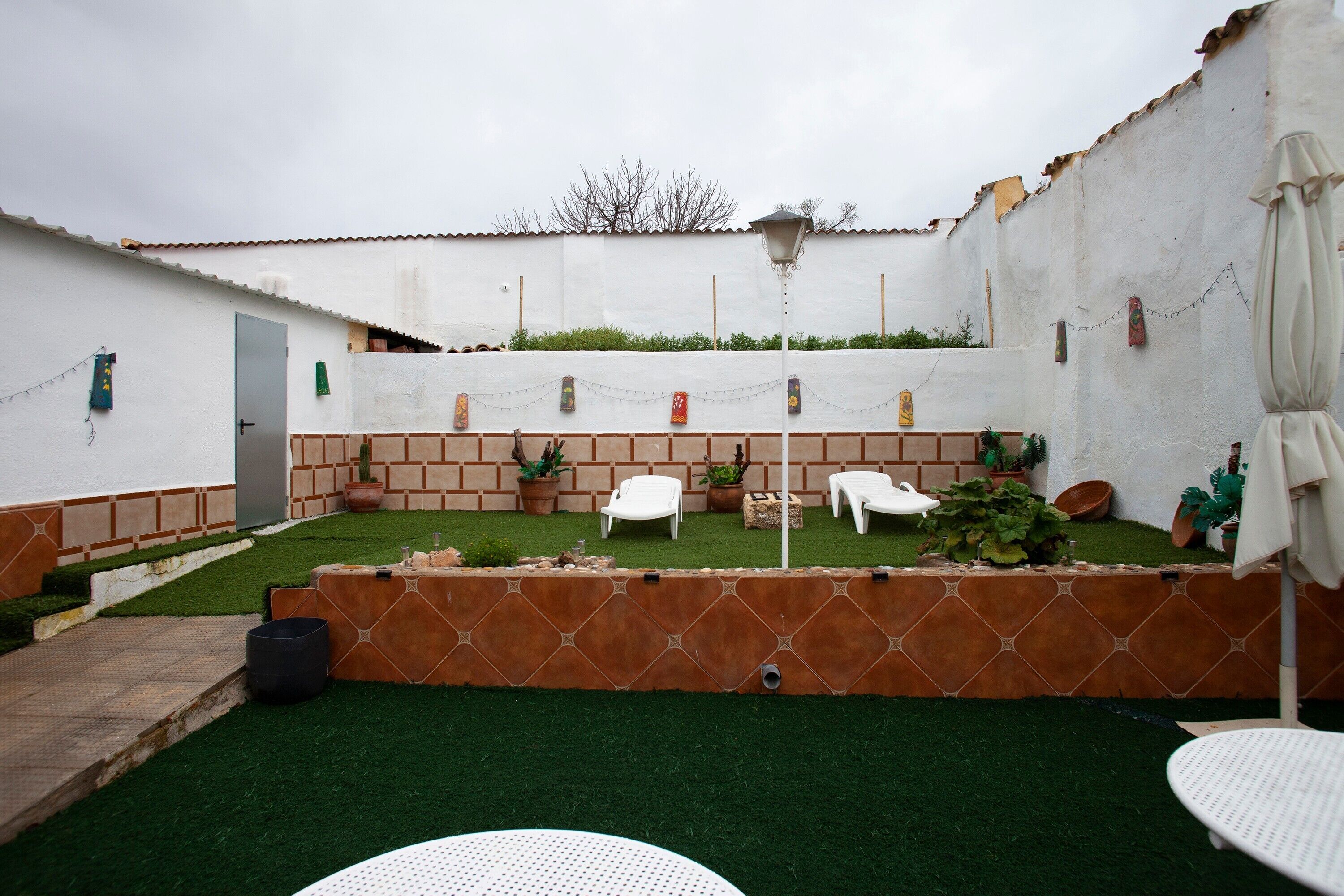 Une cour avec une pelouse artificielle, des chaises blanches, des plantes en pot et un mur blanc avec une porte.